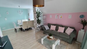 Un salon moderne avec une table basse en verre, un canapé gris agrémenté de coussins roses, un coin repas avec une table et des chaises blanches, et un espace cuisine avec une plante et des verres à vin posés sur le comptoir.
