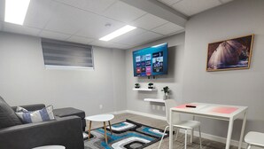 A modern living room with a grey sofa, a white coffee table, a small white table with two chairs, a blue and grey patterned rug, and a flat-screen TV mounted on the wall displaying various channels. - Spacious Basement Suite in Bonavista With Separate Entrance (Winnipeg)