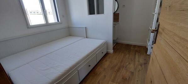 Una habitación pequeña con cama individual, piso de madera, ventana y pared blanca.