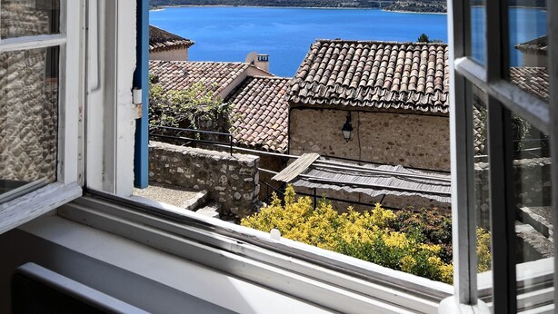 Vista dalla finestra su una cittadina costiera con edifici dalle tegole color terracotta e un cielo azzurro intenso.