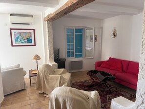 A living room with a red sofa, a covered chair, a table with a lamp, a framed picture, and a window with blue shutters.