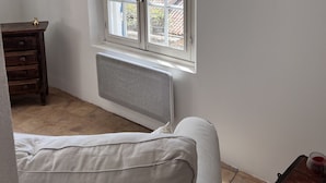 Una stanza con un'ampia finestra, un termosifone, una cassettiera in legno, un divano bianco e un piccolo tavolo con un bicchiere.