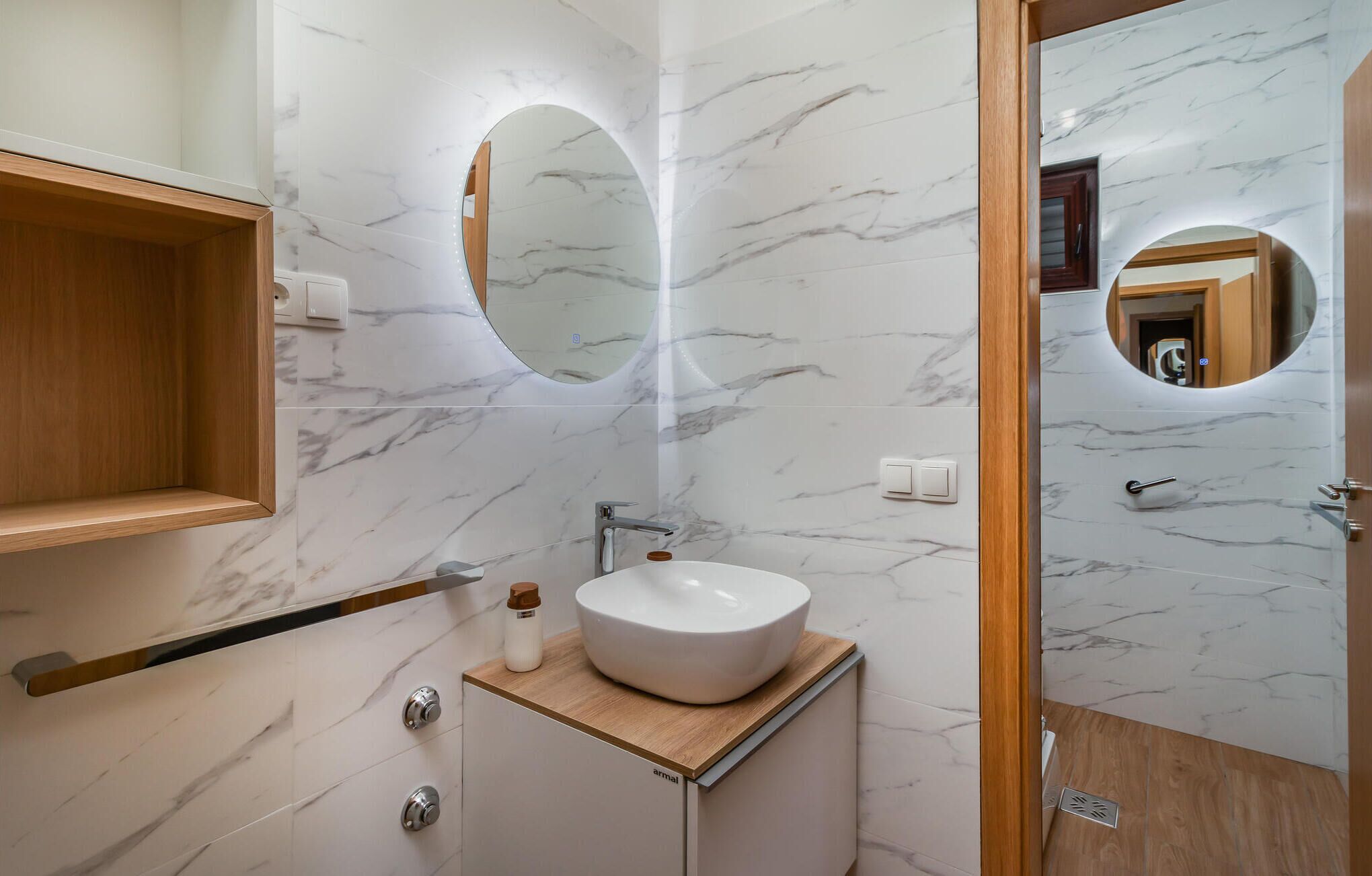 Un baño moderno con salpicadero de mármol, tocador de madera, espejo redondo y lavabo blanco.