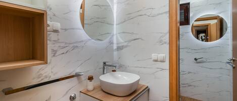 Un baño moderno con salpicadero de mármol, tocador de madera, espejo redondo y lavabo blanco.