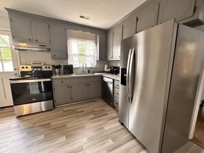 A modern kitchen with stainless steel appliances, light wood flooring, and a window with patterned curtains. - Pet Friendly Cozy & Comfy 3 BR House w Large Yard & Deck (Mechanicsville)