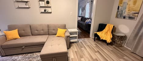 A living room with a sectional sofa, ottoman, and a small side table. There are wall-mounted shelves with decorative items and a painting on the wall.