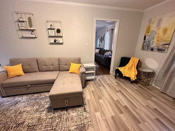 Ein Wohnzimmer mit Ecksofa, Hocker und einem kleinen Beistelltisch. An den Wänden befinden sich Regale mit Dekoration und ein Gemälde.