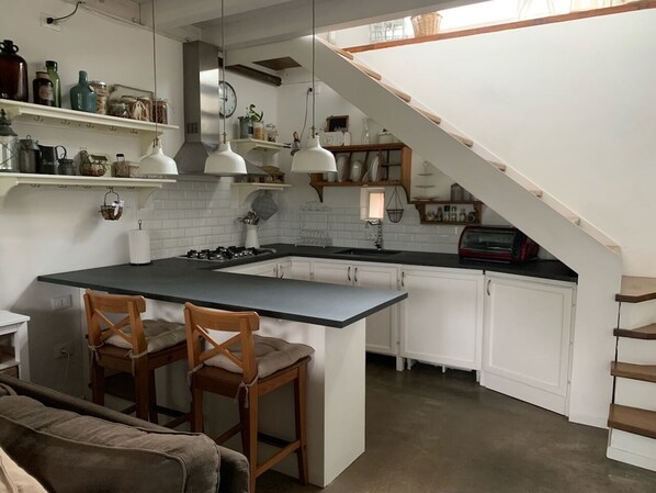 A kitchen with white cabinets, a central island with two chairs, pendant lights, and a staircase leading to a loft area. - The little house in Verciano (Capannori)