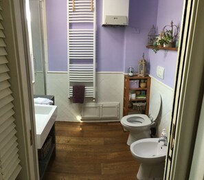A bathroom with a bathtub, toilet, and sink. There is a wooden shelf with decorative items and a towel hanging on the wall. - The little house in Verciano (Capannori)