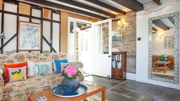 A cozy living room with a floral sofa, a coffee table with books, a stone wall, and a mirror with a patterned frame.
