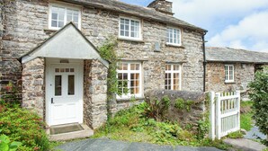 A stone house with a white door, multiple windows, and a small garden area with plants and a white fence.