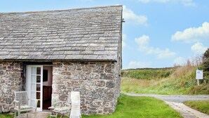 A stone building with a door leading inside, a small outdoor seating area with a chair and table, and a grassy area with a path.