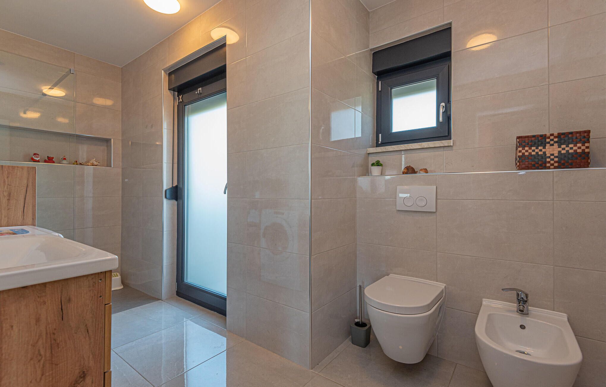 Un bany modern amb vàter, bidet i rentapel. Hi ha un armari de fusta i una petita estant amb objectes decoratius.
