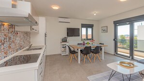 Ett modernt kök med kakelbeklädd vägg bakom arbetsbänken, en matplats med bord och stolar, en TV som är monterad på väggen och en balkong med utsikt.