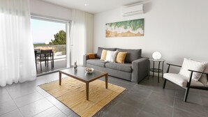 Uma sala de estar moderna com um sofá cinzento, uma mesa de centro e uma poltrona. Há uma janela grande com cortinas, um quadro na parede e um aparelho de ar condicionado.