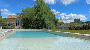 A clear swimming pool with a tiled deck, surrounded by greenery and a house with a tiled roof.