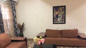A living room with a brown sofa, a black coffee table with a vase of flowers, and a painting on the wall. - Dazzle Hotels and Apartments (Abuja)