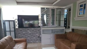 A living room with leather sofas, a TV mounted on a stone wall, a kitchen area with cabinets and a glass door, and a view of the outside. - Dazzle Hotels and Apartments (Abuja)