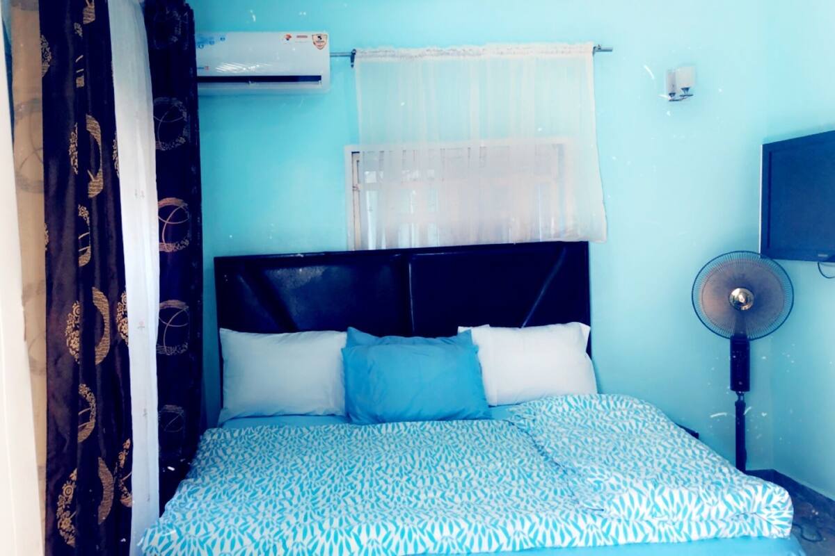 Un dormitorio con una cama, una ventana con cortina translúcida, un aire acondicionado fijado a la pared, un ventilador de pie y un televisor en la pared.