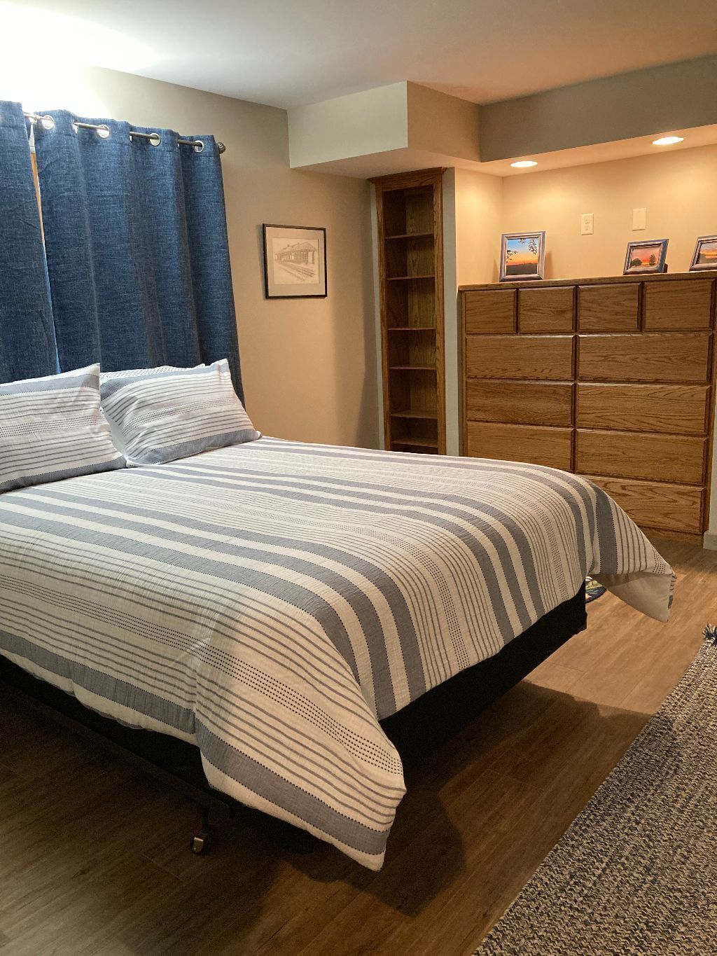 Une chambre à coucher avec un lit, une armoire en bois, un tableau encadré au mur et un rideau bleu.