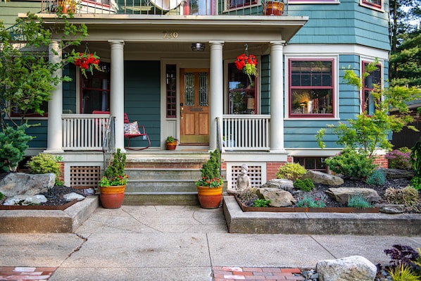 Une maison à deux étages avec un porche, située au numéro 230, et dotée d’un jardin bien entretenu avec des plantes en pot et une statue.