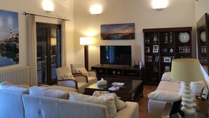 Un salon comprenant un canapé, des fauteuils, une table basse, un téléviseur, une lampe, une bibliothèque et un tableau.