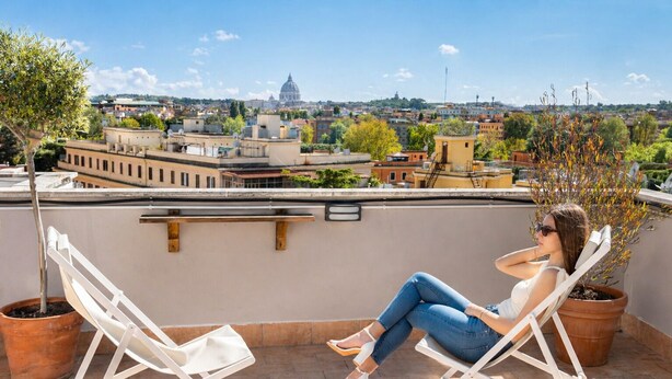 A woman sitting on a white lounge chair on a rooftop terrace with potted plants and a cityscape view in the background.