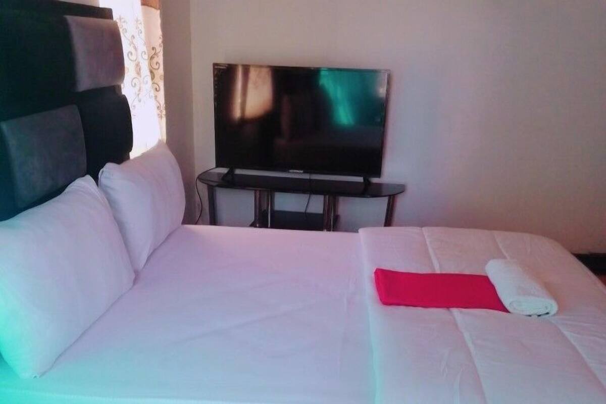 Ein ordentlich bezogenes Bett mit weißen Laken und einer roten Decke, ein Fernseher auf einem Schrank und ein Fenster mit Vorhängen.