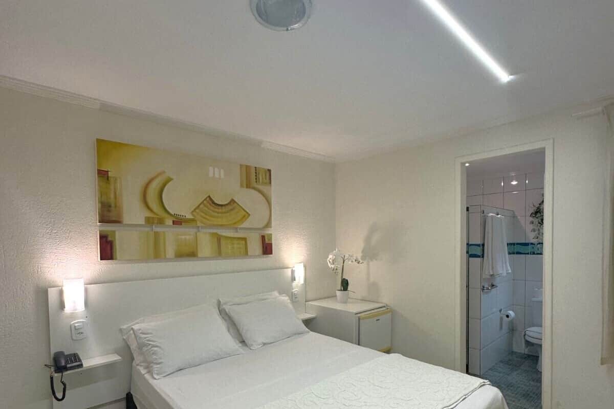 A hotel room with a bed, bedside tables, a nightstand, a wall-mounted lamp, and a shower area.