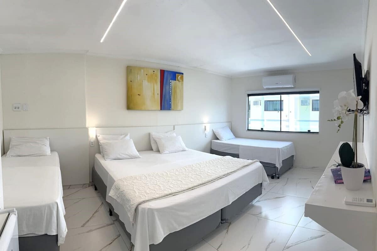A modern hotel room with a large bed, two smaller beds, a desk with a computer, and a large window with a view.