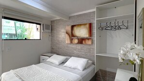 A modern bedroom with a bed, a nightstand, a built-in wardrobe, and a window with a view of greenery. - POUSADA OCEANIC (Arraial do Cabo)