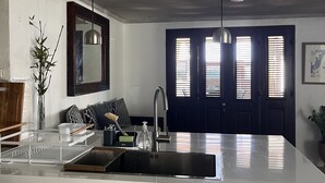 A modern kitchen with a white countertop, stainless steel sink, and white cabinets. There is a dark blue door with windows, a hanging pendant light, and a vase with green plants on the left side. - Beachfront 2 bedroom apartment with private pool -NEW LISTING (Fitts Village)