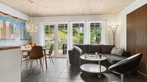 Ein modernes Wohnzimmer mit einem dunkelgrauen Sofa, einem runden Couchtisch und einem Essbereich mit einem weißen Tisch und Stühlen. Große Fenster bieten Blick auf eine grüne Landschaft.