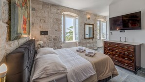 Une chambre avec un mur en pierre, un lit avec du linge de lit blanc, une commode, un téléviseur et une fenêtre donnant sur un beau paysage.