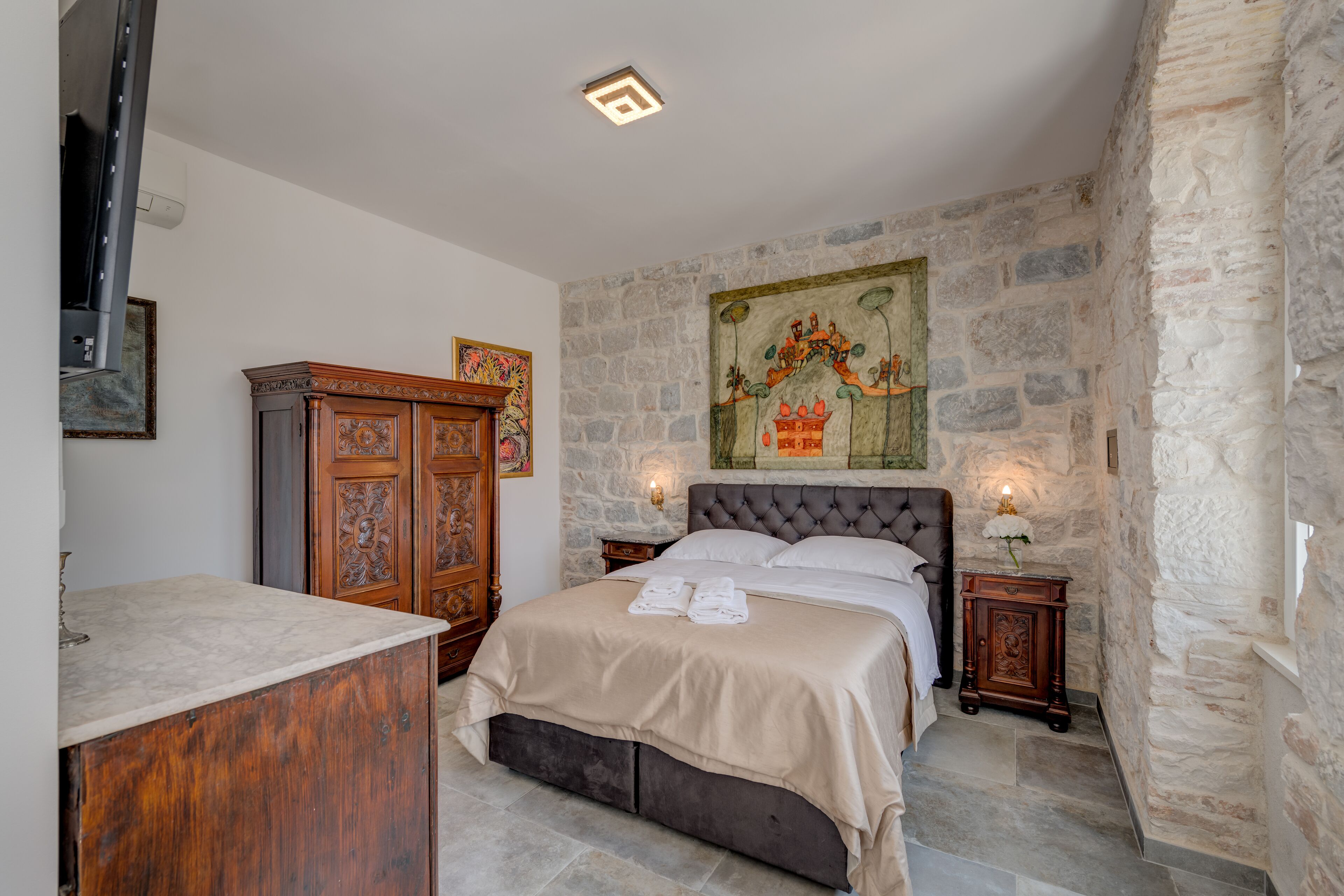 A bedroom with a stone wall, a bed with a grey headboard, a wooden cabinet, and a painting on the wall.
