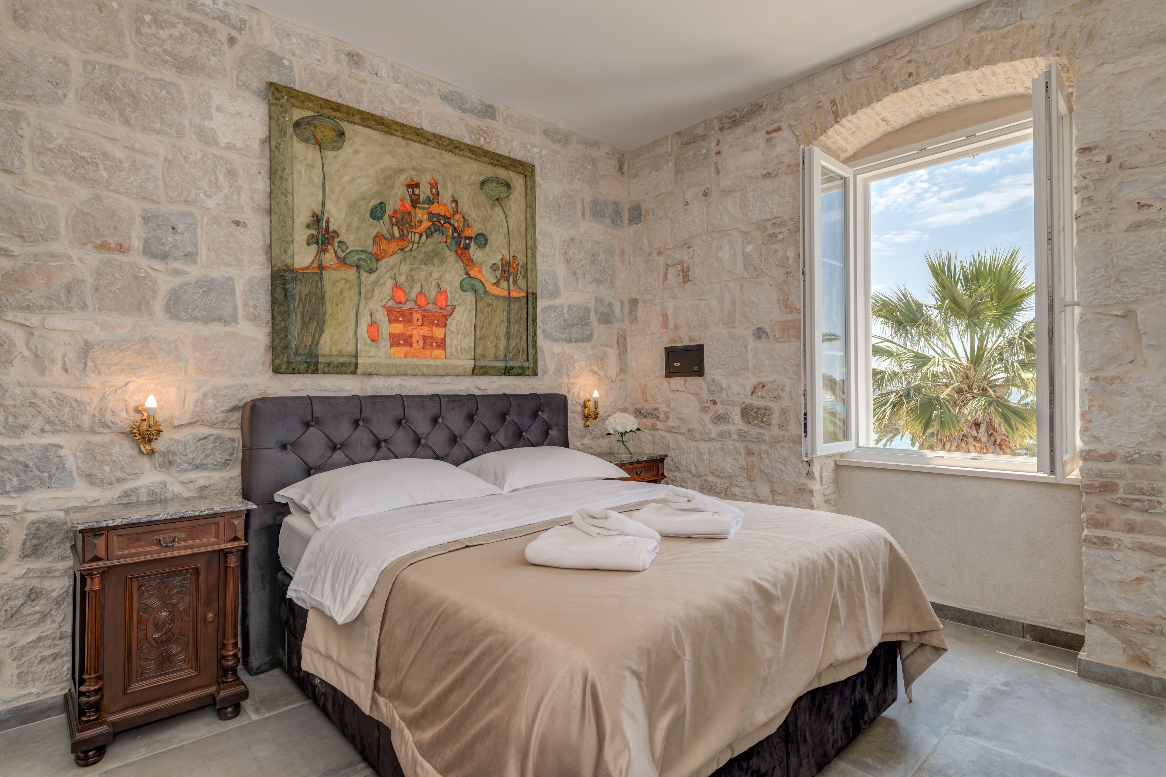A bedroom with a stone wall, a large bed, a painting, and a window with a view of palm trees.