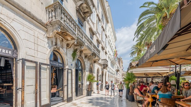 Une rue bordée de terrasses, de palmiers et d’un immeuble abritant des boutiques.