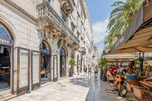 Calle con mesas al aire libre, palmeras y un edificio con tiendas.