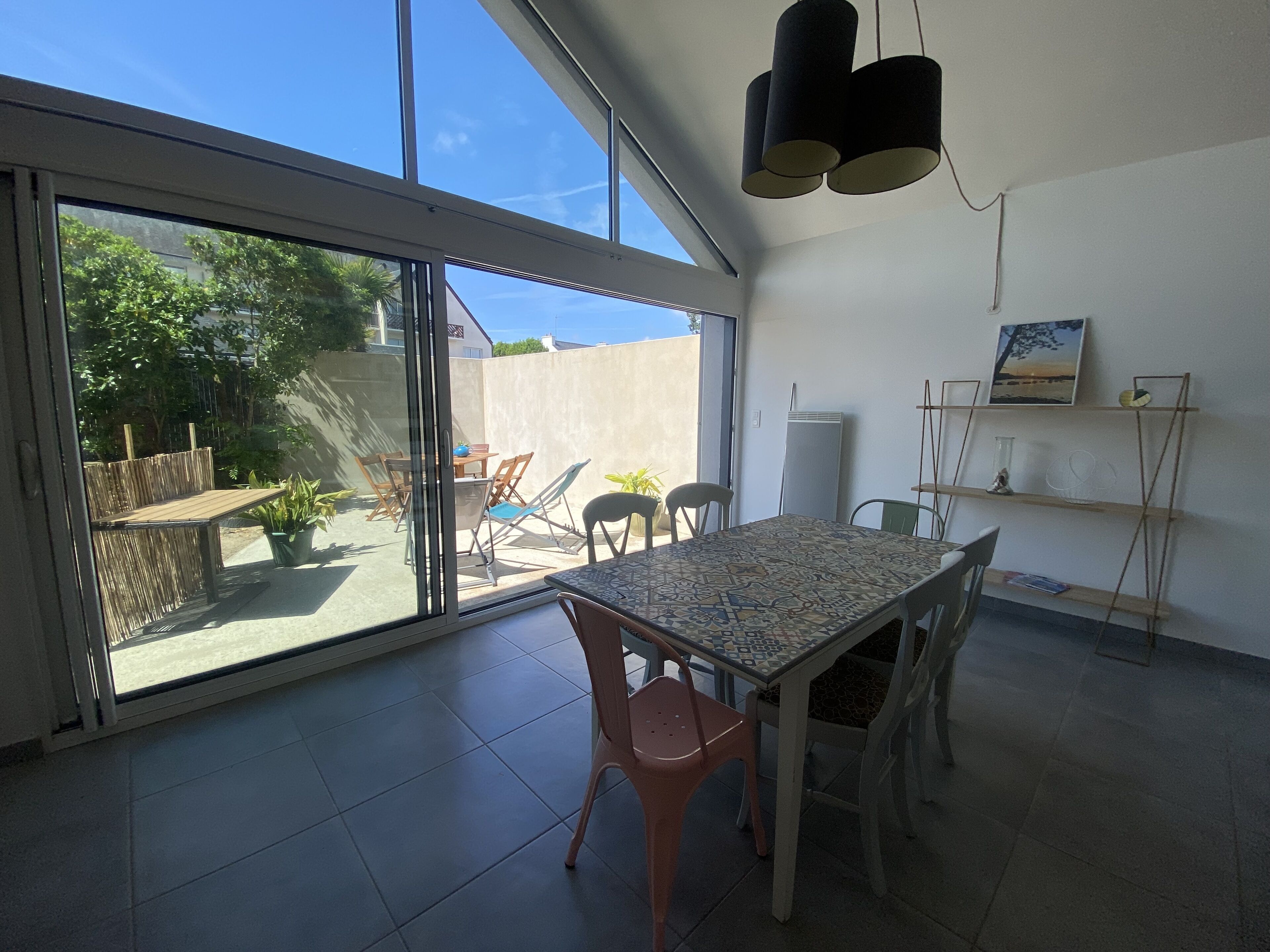 Une pièce dotée d’une grande porte-fenêtre coulissante donnant sur un patio extérieur. À l’intérieur, on trouve une table avec des chaises et un luminaire fixé au plafond.