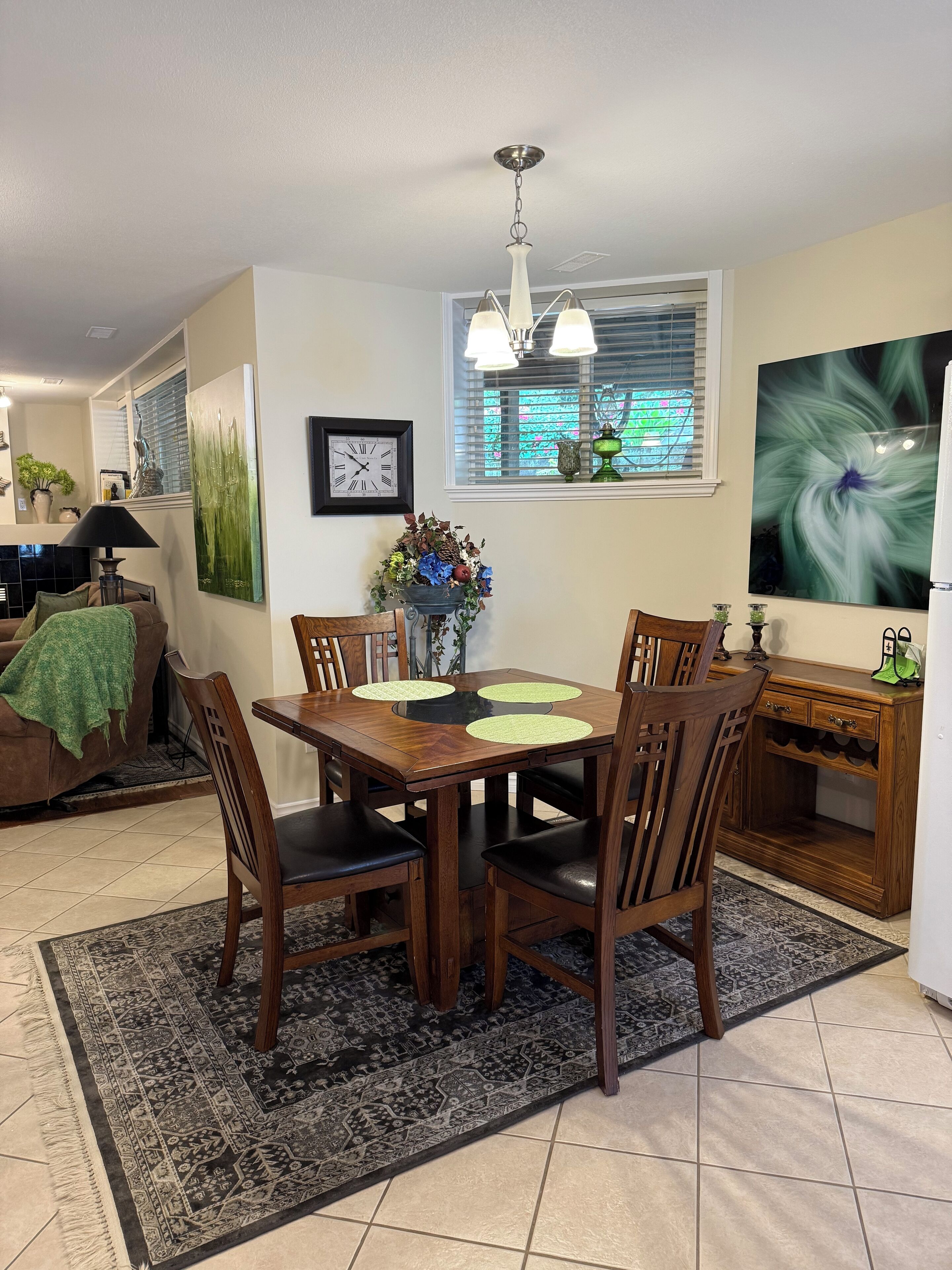Un coin repas avec une table et des chaises en bois, une horloge au mur et une fenêtre avec des stores.