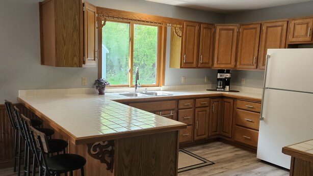 A kitchen with wooden cabinets, a large island with bar stools, a refrigerator, and a window with a view of greenery.