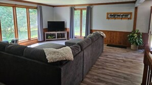 A living room with a sectional sofa, a flat-screen TV, and large windows.