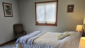 A bedroom with a bed, a chair, a nightstand with a lamp, and a window with blinds.