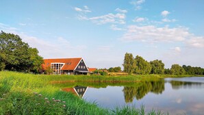 A traditional Dutch farm house with a red roof, situated next to a calm lake surrounded by lush greenery and trees. - Nice home in Heinkenszand with WiFi (Heinkenszand)