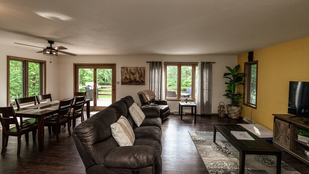A living room with a brown leather sofa, dining table, and chairs. There is a ceiling fan, large windows, and a TV on a wooden cabinet. - Whispering Pines By The Bay, quiet seclusion near water and town (Sturgeon Bay)