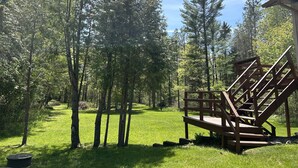 A wooden deck with a staircase leading to a building, surrounded by a grassy area and tall trees. - Whispering Pines By The Bay, quiet seclusion near water and town (Sturgeon Bay)