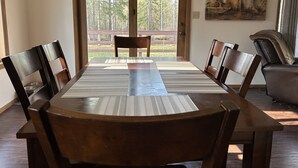 A dining room with a large table and chairs, a painting on the wall, and a view of trees through a glass door. - Whispering Pines By The Bay, quiet seclusion near water and town (Sturgeon Bay)