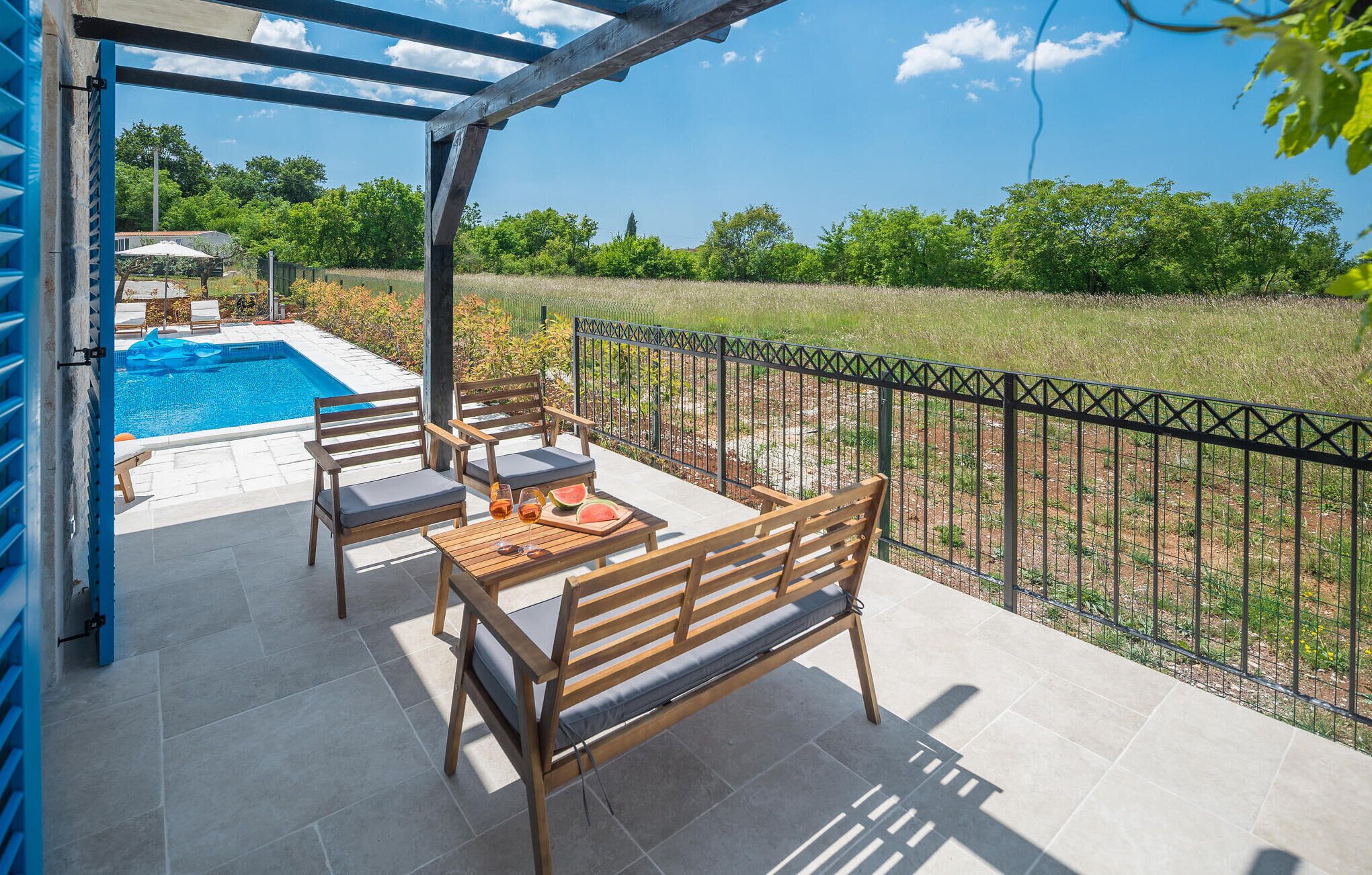 An outdoor patio with a pool, wooden furniture, and a view of a grassy area.