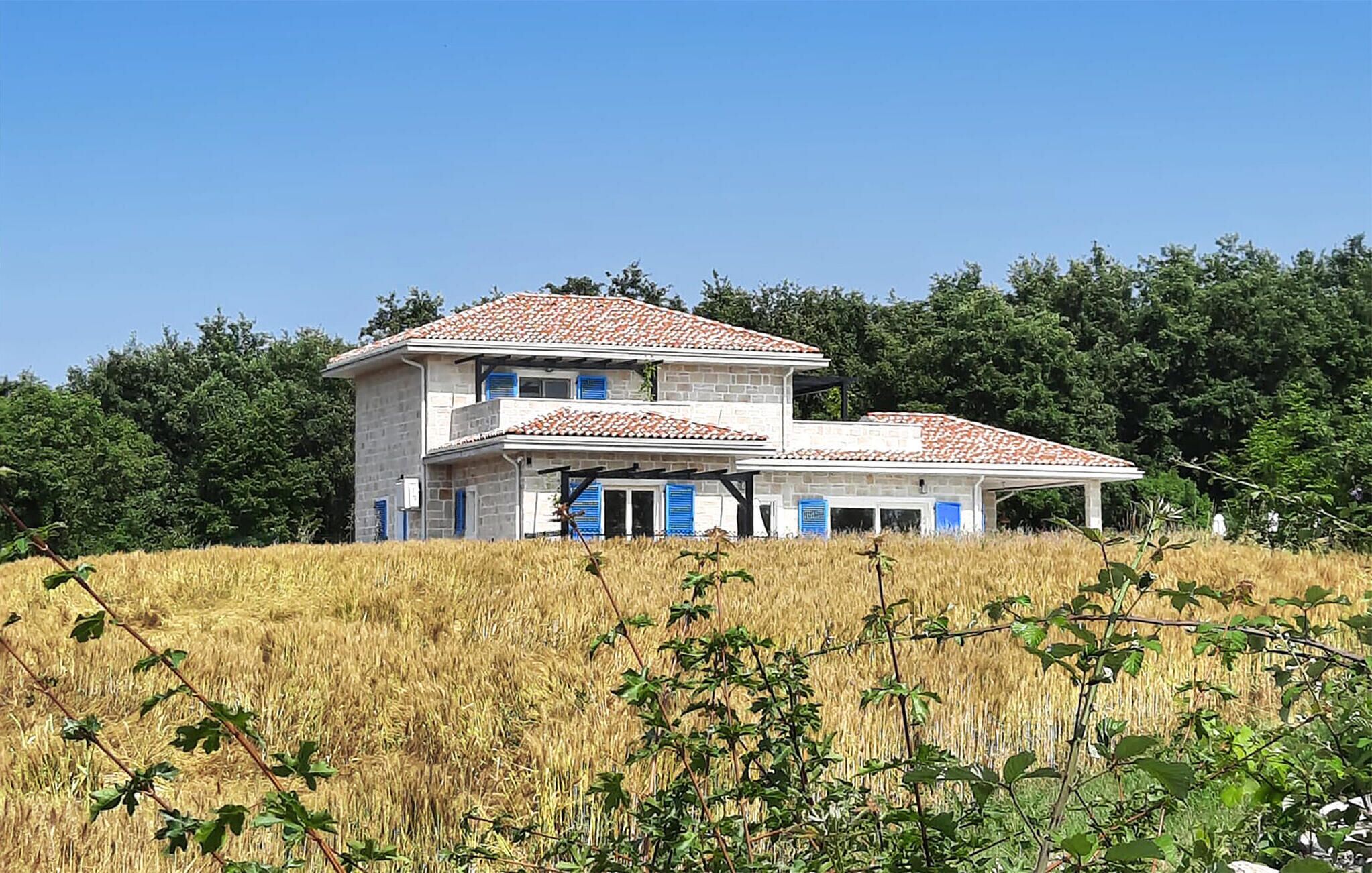 Une maison à deux étages, avec un toit recouvert de tuiles, entourée d’un champ de blé et bordée d’arbres au loin.