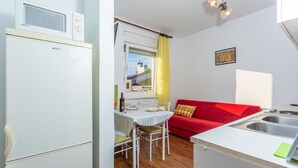A compact kitchen with a refrigerator, a dining table with chairs, a red sofa, and a window with a yellow curtain.